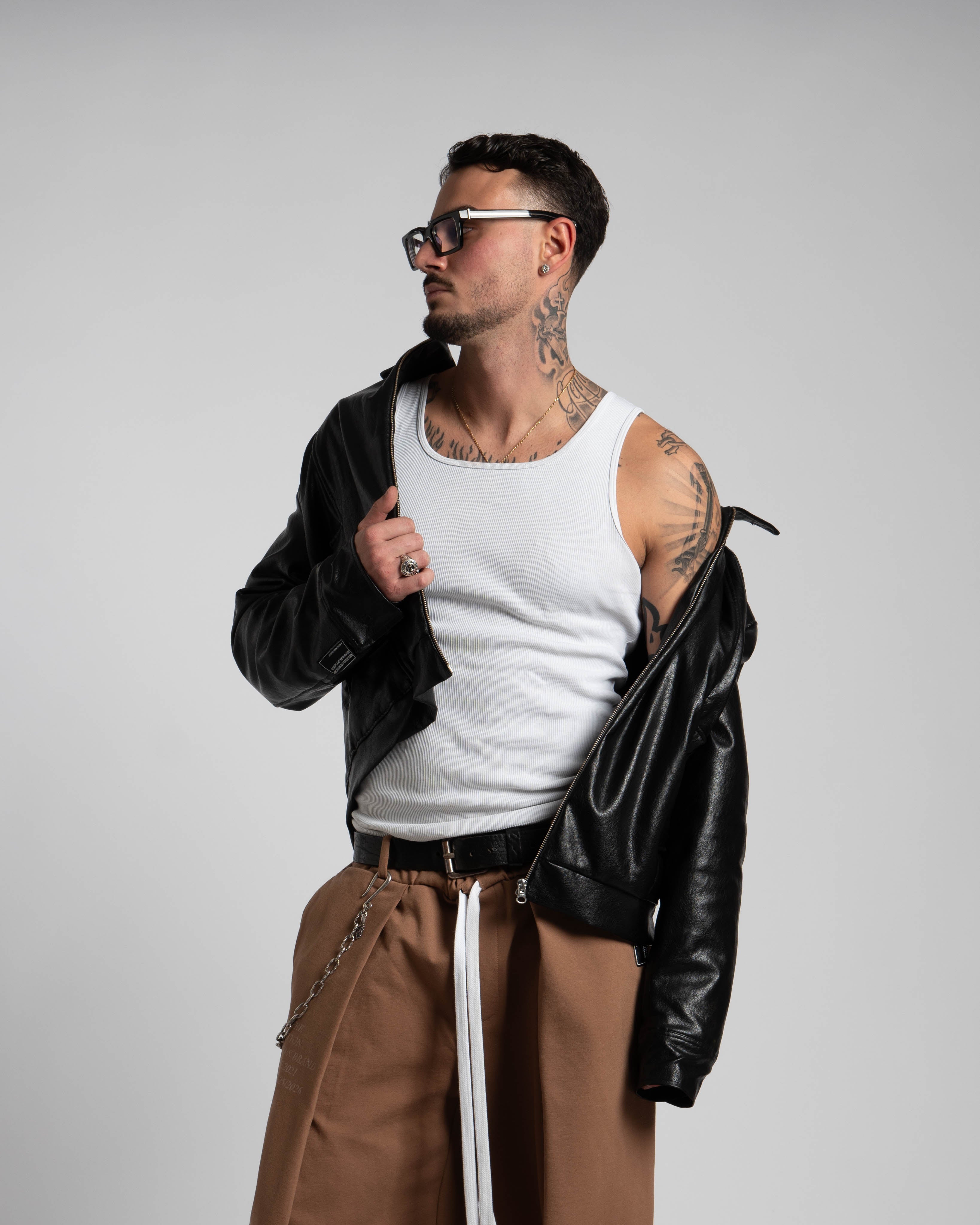 Man wearing a white tank top, brown pants, and a black leather jacket on a plain background