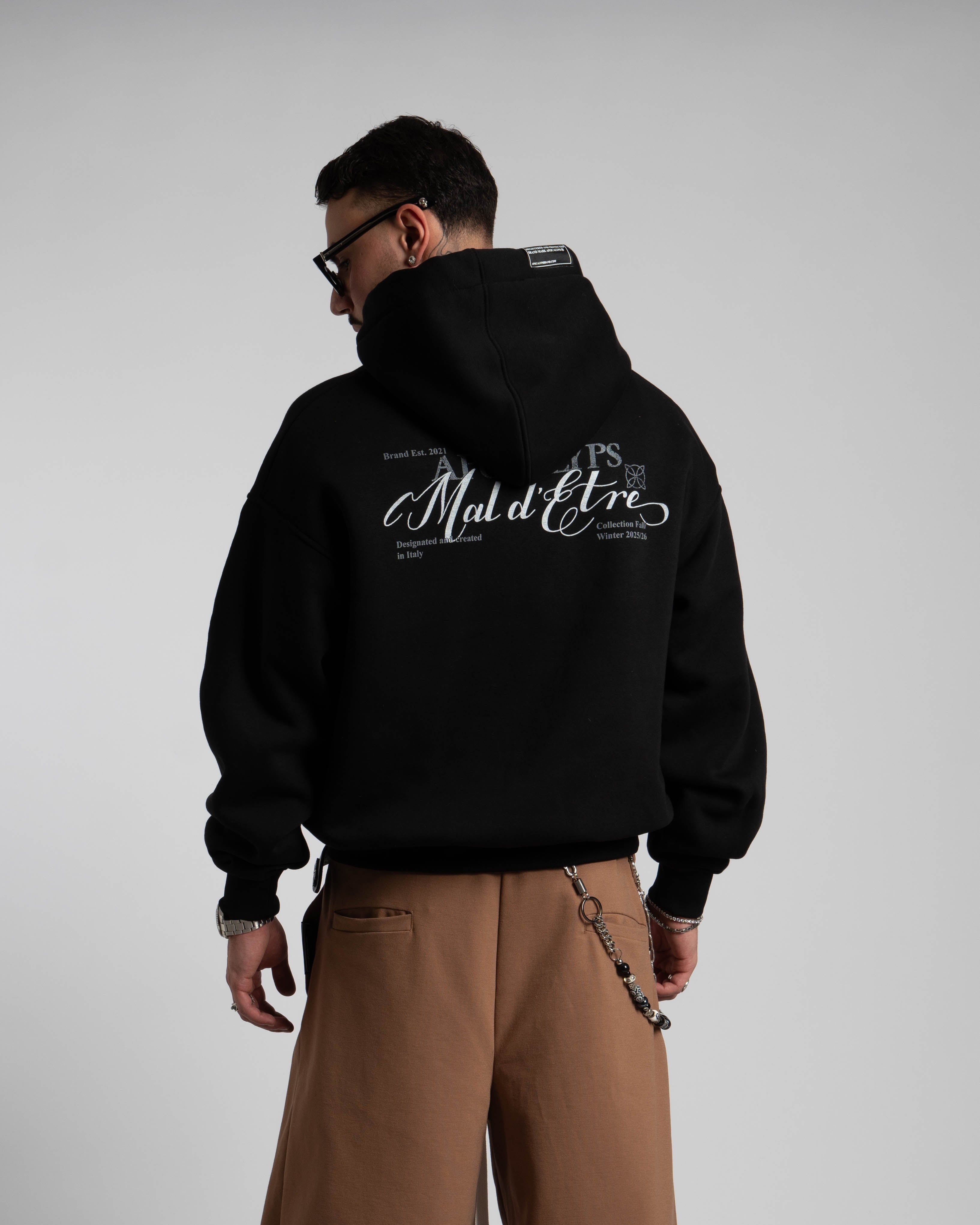 Person wearing a black hoodie with text on the back and brown pants, standing against a plain background.