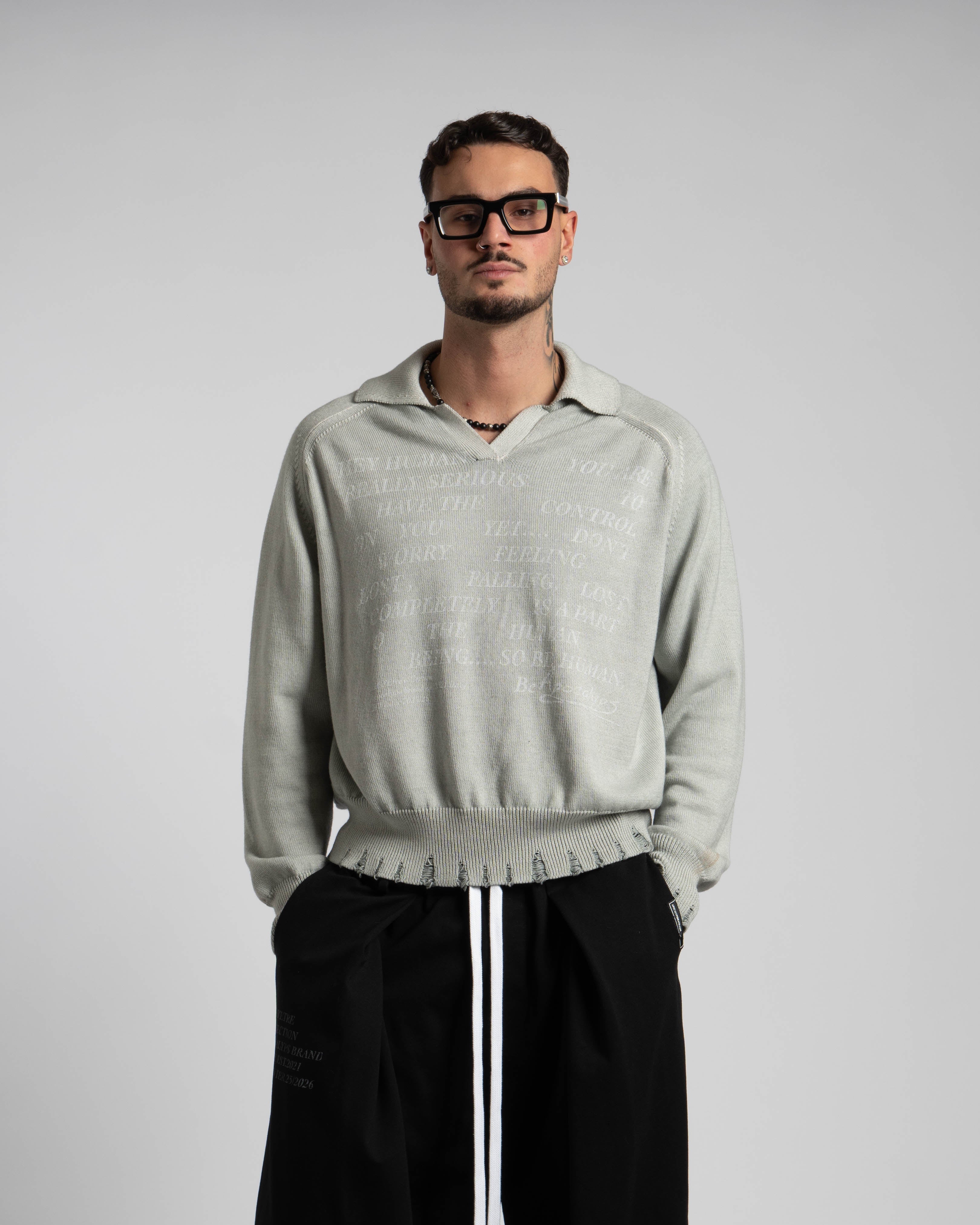 Man wearing a light gray sweater and black pants against a plain background